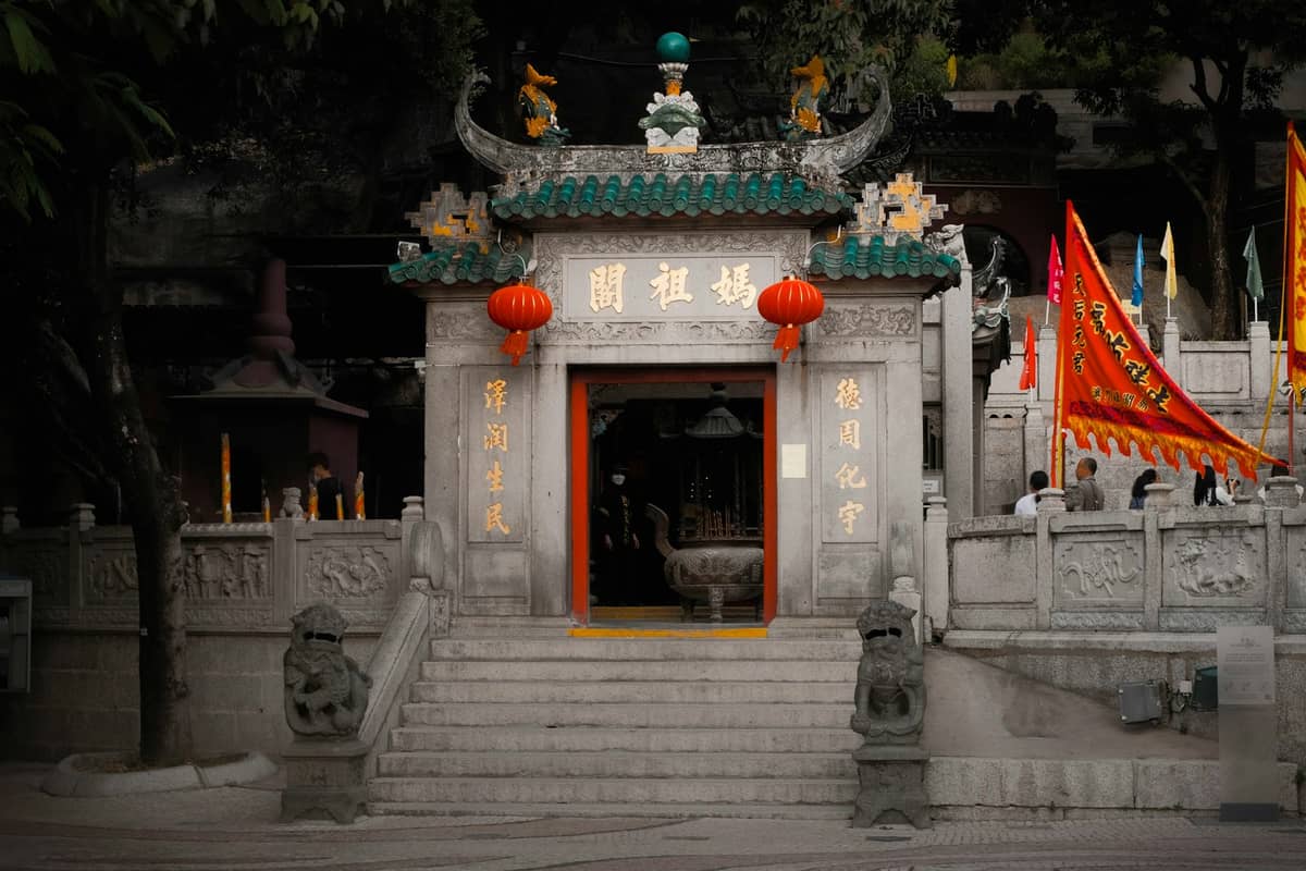 Eingang zu einem prunkvollen chinesischen Tempel in Macau mit Steinstufen, grünem Ziegeldach, roten Laternen und gemeißelten Steinlöwen zu beiden Seiten des Eingangs.