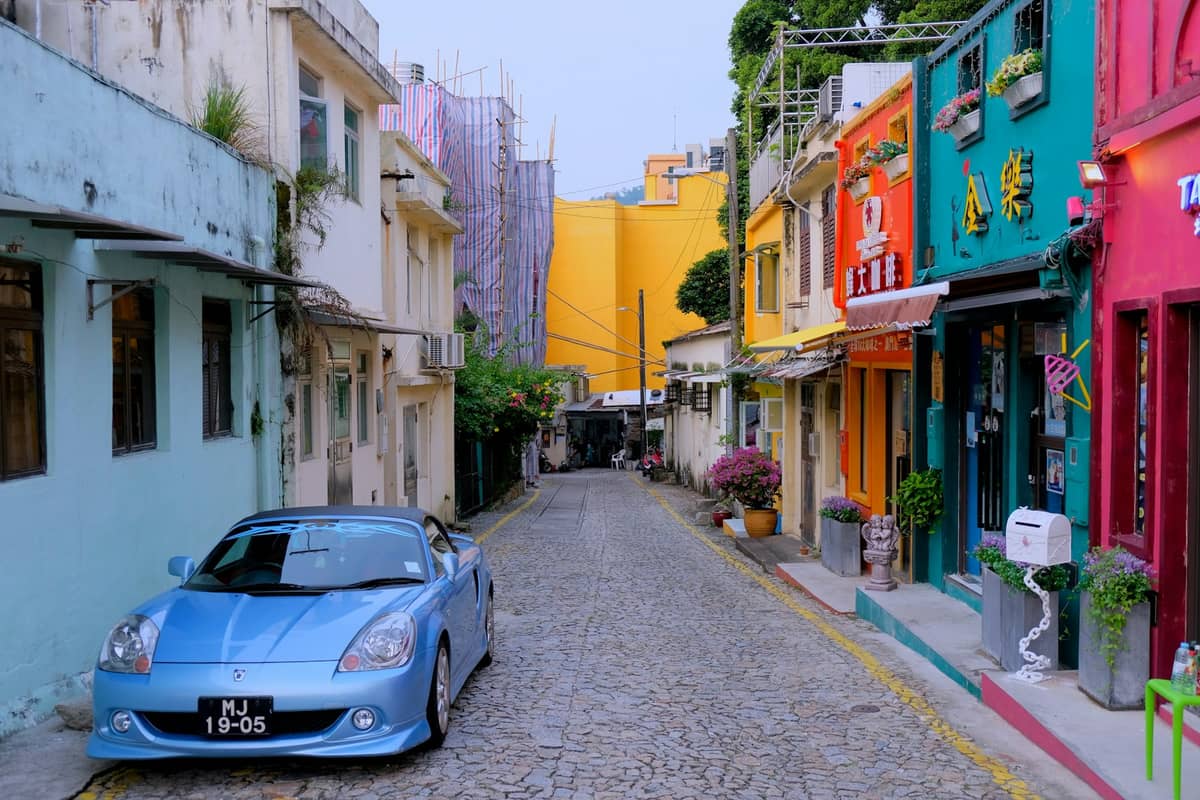 Pastellfarbene Dorfstraße in Coloane, Macau, mit traditionellen Häusern, Geschäften und einem geparkten blauen Auto auf einer ruhigen Kopfsteinpflasterstraße.