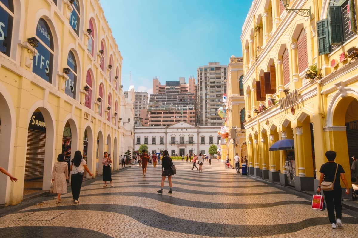 Menschen schlendern über den farbenfrohen Senado-Platz, Macaus historisches UNESCO-Stadtzentrum, umgeben von pastellfarbenen Kolonialgebäuden unter strahlend blauem Himmel.