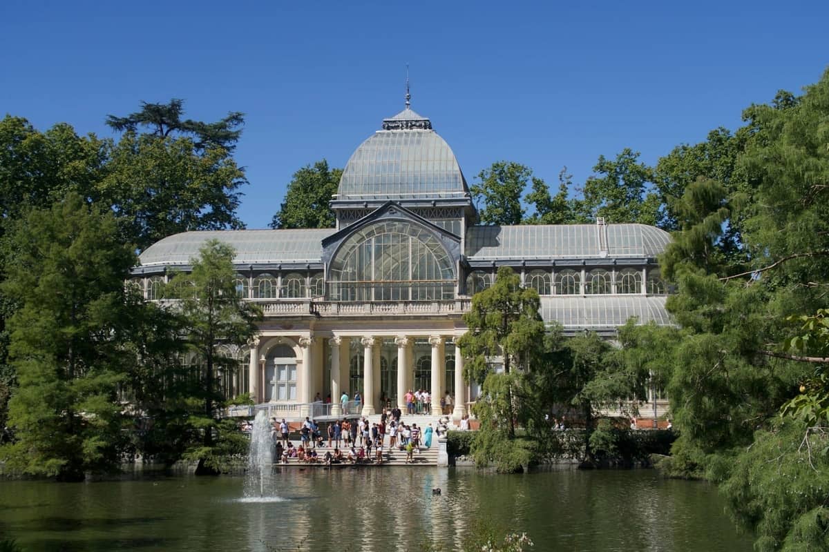 Widok na Palacio de Cristal w Madrycie z drugiego brzegu stawu, otoczony bujną zielenią i odwiedzającymi cieszącymi się parkiem.