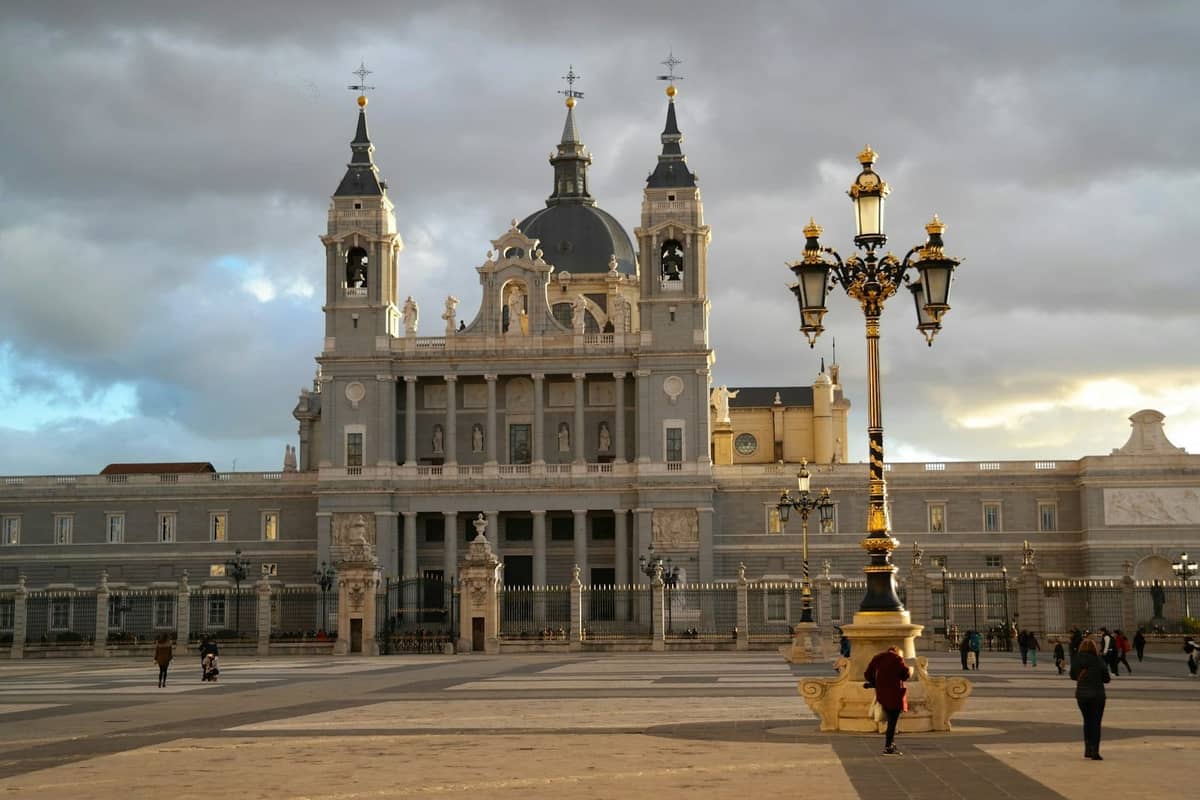 Szeroki widok na Catedral de la Almudena w Madrycie z wieżami i centralną kopułą, widziany z placu z ozdobnymi latarniami i przechadzającymi się ludźmi.