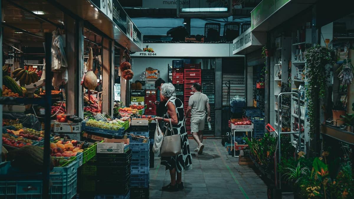 Suasana pasar dalam ruangan dengan buah dan sayuran berwarna-warni, para pembeli yang menjelajahi lapak, dan produk lokal alami dalam suasana yang ramai.