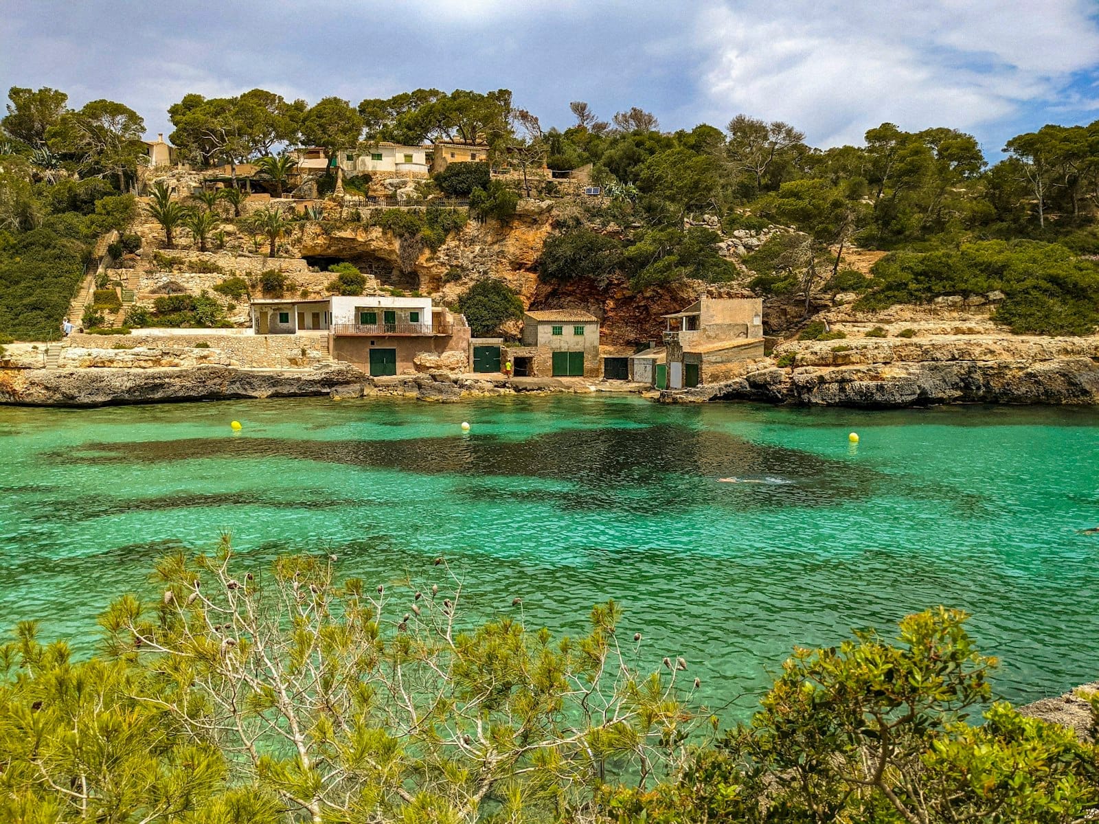 Enseada em Maiorca com águas turquesa, casinhas de pescadores nas falésias rochosas e vegetação exuberante sob um céu azul.