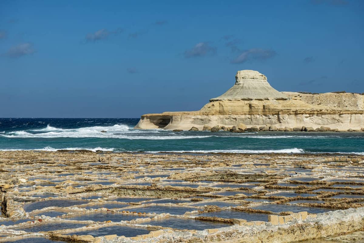 Wybrzeże Gozo z charakterystycznymi słonymi jeziorami na pierwszym planie i wyróżniającym się skalistym wzgórzem nad Morzem Śródziemnym pod bezchmurnym niebieskim niebem.