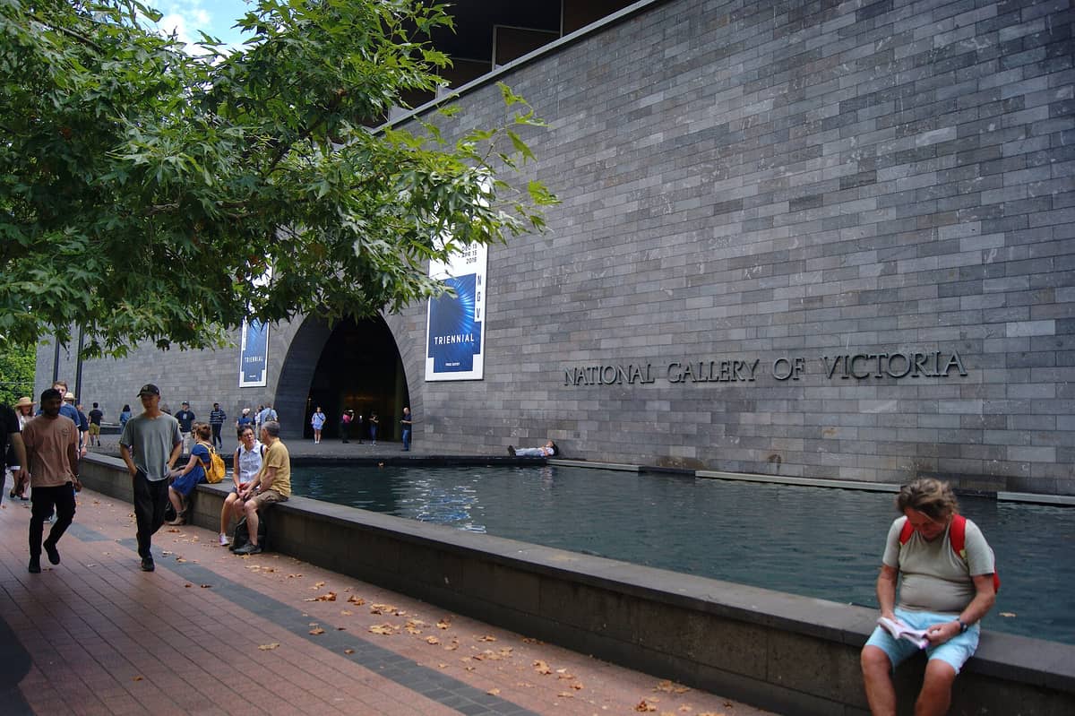 Des personnes assises et marchant devant la National Gallery of Victoria, un grand musée d'art gratuit à Melbourne avec une façade moderne.