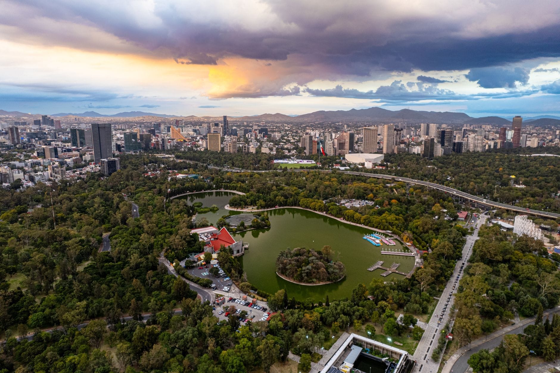 Vista aérea do Parque Chapultepec na Cidade do México com exuberantes áreas verdes, lagos, pedalinhos e o horizonte da cidade ao pôr do sol.