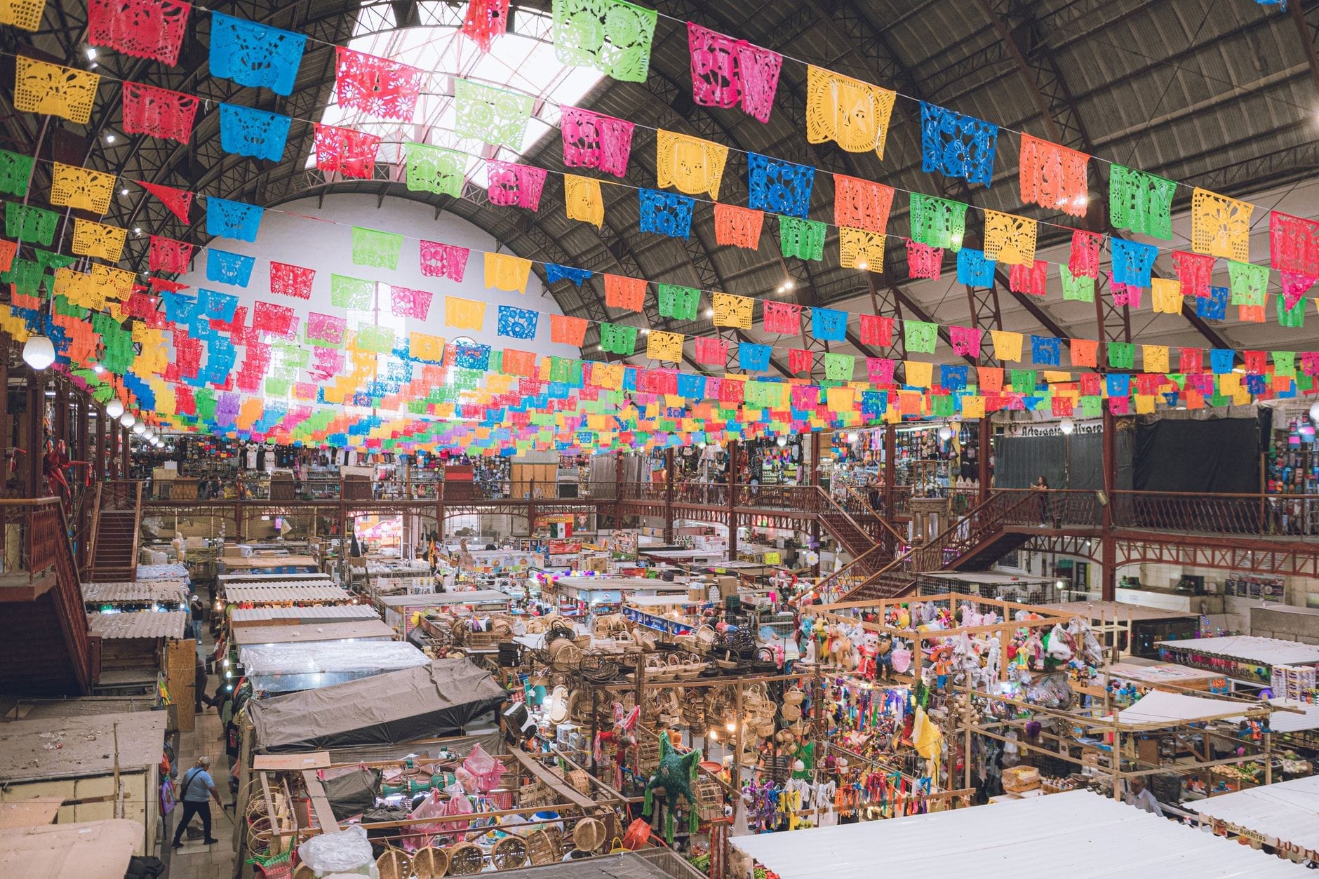 Cena animada de mercado mexicano em ambiente interno bem iluminado com coloridas bandeirolas de papel picado acima e barracas de vendedores movimentadas abaixo.
