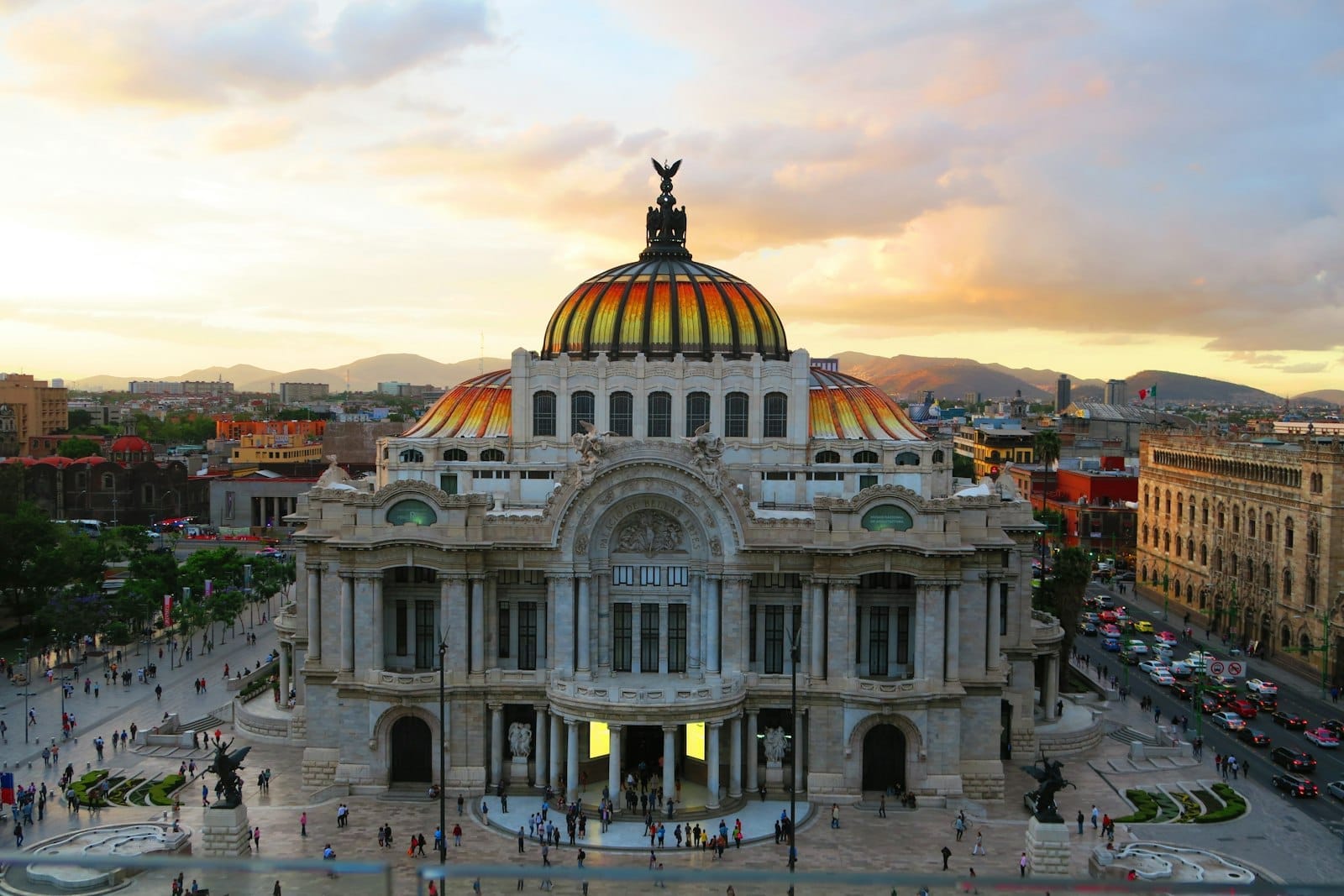 Palácio de Belas Artes da Cidade do México ao entardecer, com multidão na praça, montanhas ao fundo e céu com cores vibrantes.