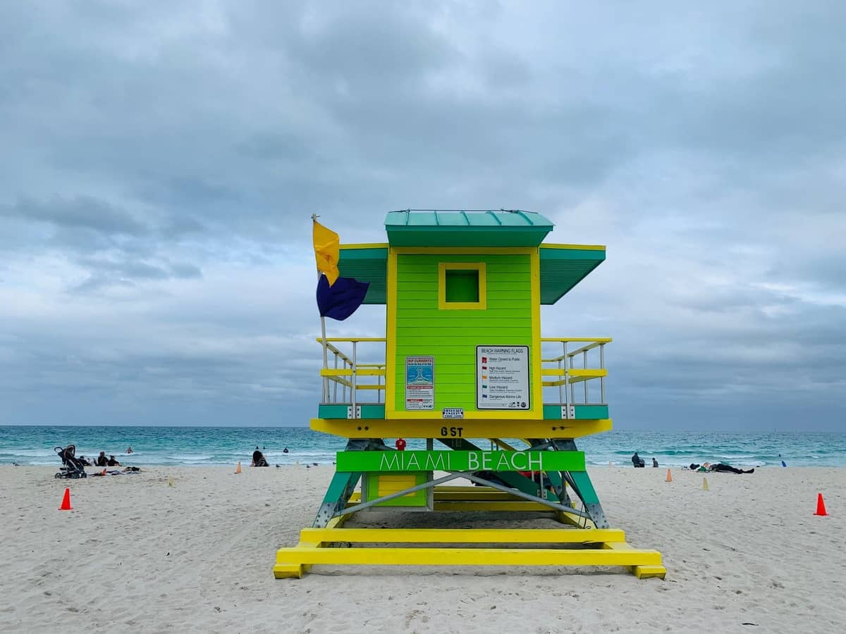 A colorful green and yellow lifeguard tower on Miami Beach with people relaxing on the white sand and turquoise ocean in the background.