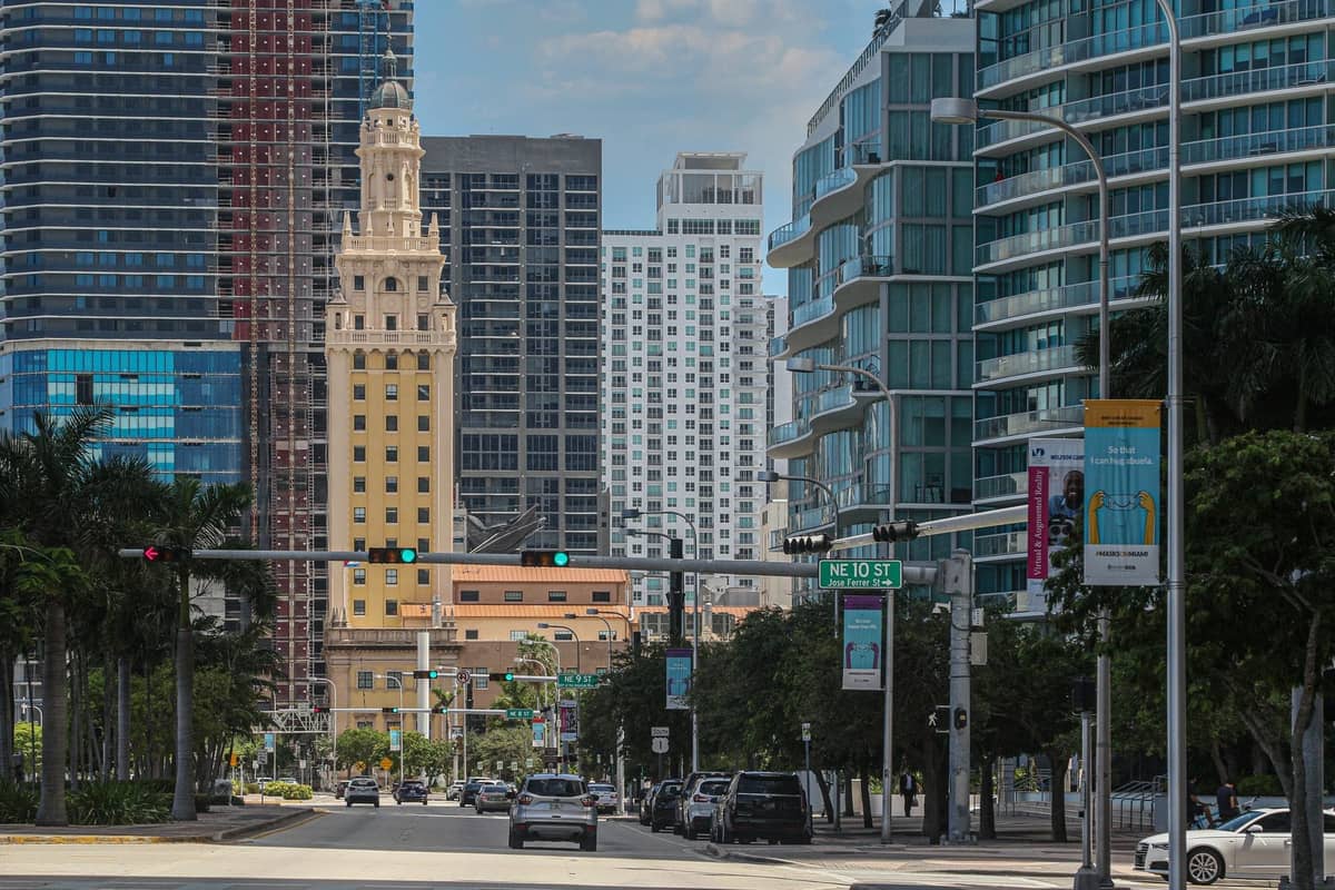 Wide street view with Miami's Freedom Tower in the center, surrounded by modern downtown skyscrapers and palm trees under a blue sky.