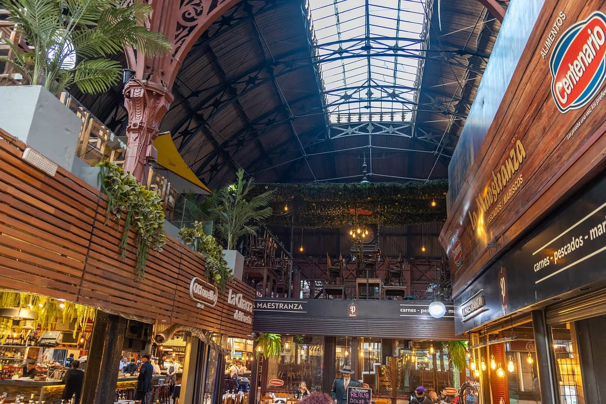 ภายใน Mercado del Puerto ในมอนเตวิเดโอ พร้อมคานไม้ แผงตลาด และผู้คนที่รับประทานอาหารใต้หลังคาเหล็กและกระจกสูง