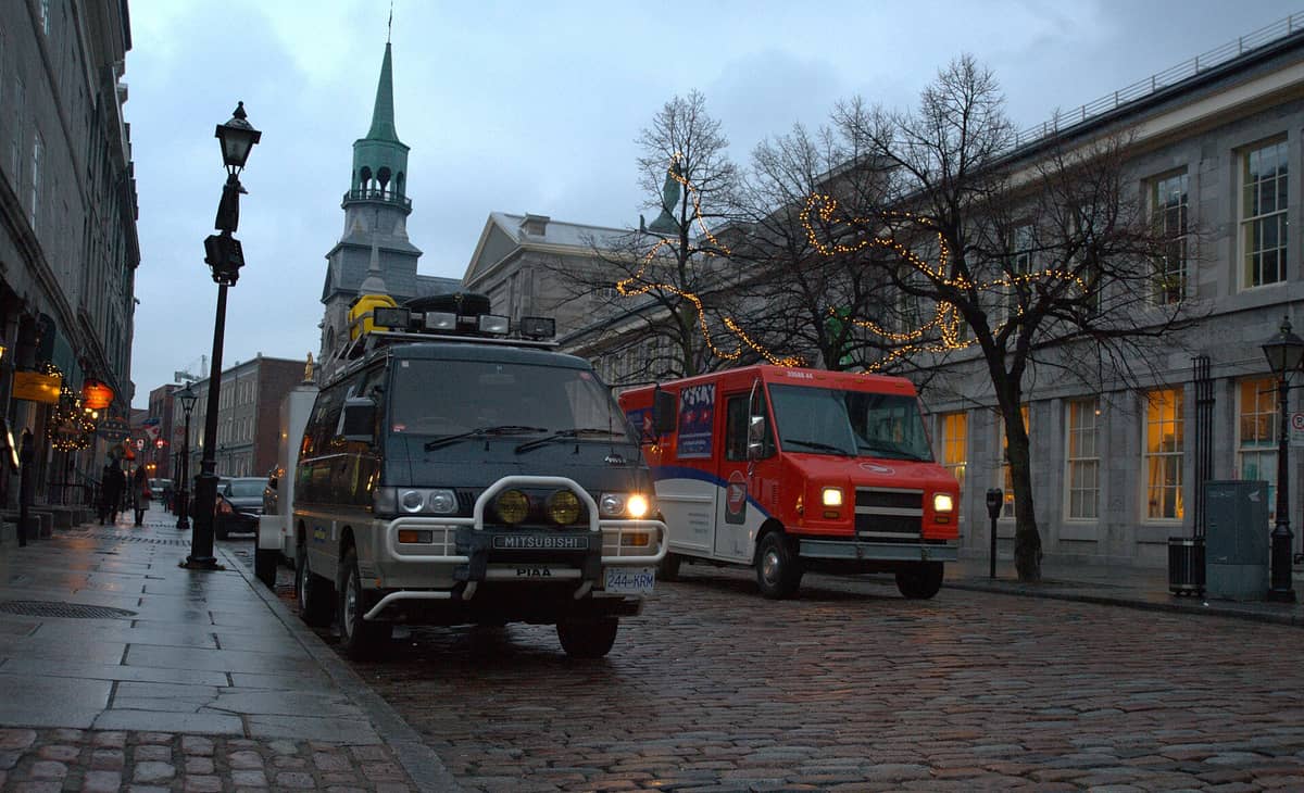 วิวถนนหินกรวดใน Vieux-Montréal พร้อมอาคารหินประวัติศาสตร์ ยอดโบสถ์ ไฟประดับ และยานพาหนะที่จอดอยู่ริมทาง