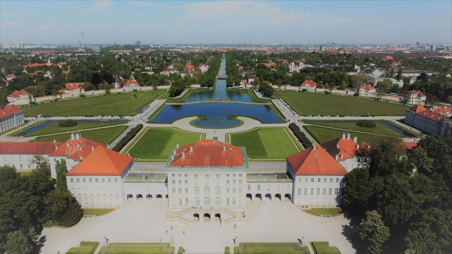 มุมมองทางอากาศของพระราชวัง Nymphenburg ในมิวนิก พร้อมสวนแบบฟอร์มัล สระสะท้อนน้ำ และทิวทัศน์เมืองในพื้นหลัง
