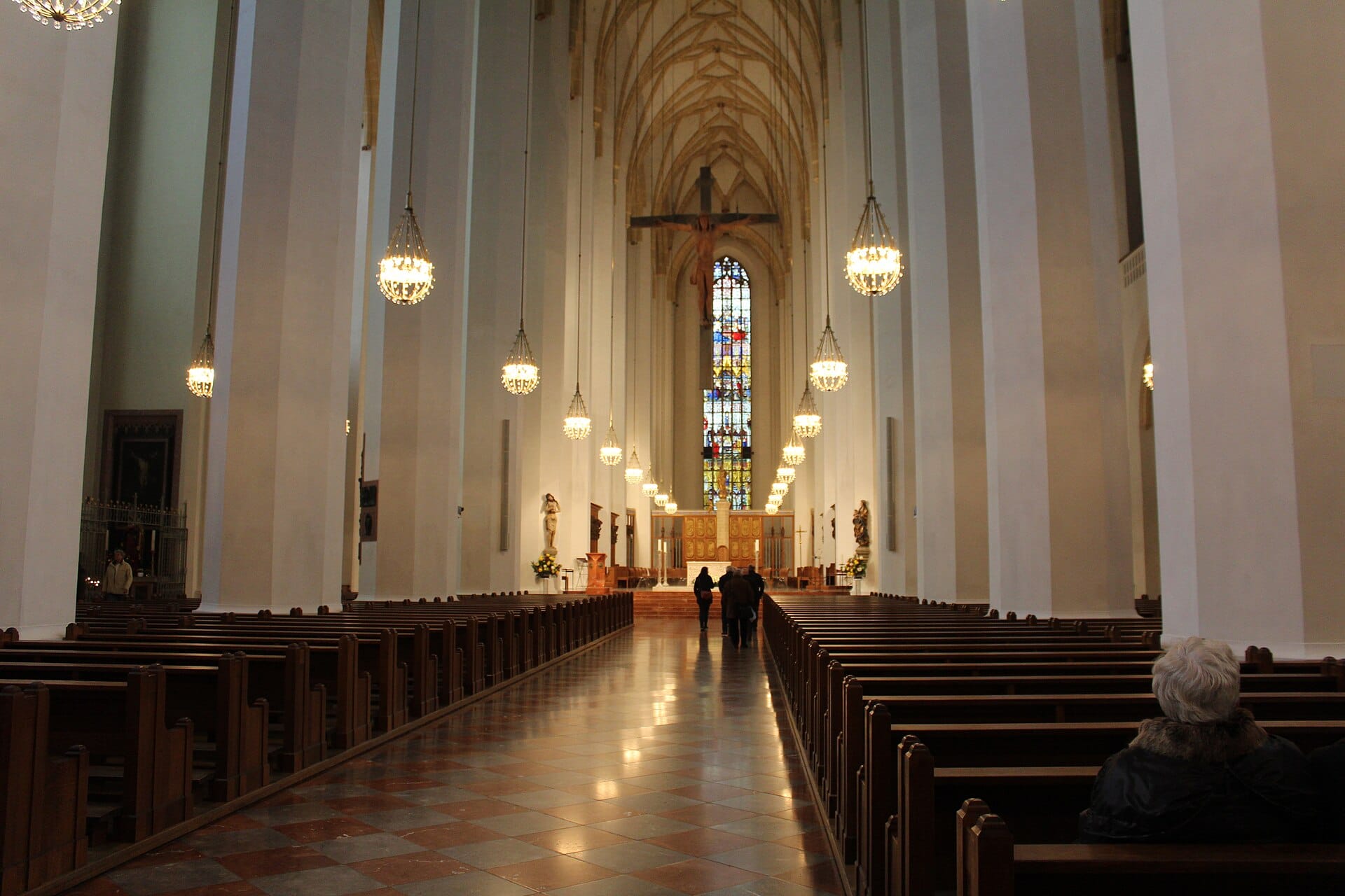ภายในโบสถ์ Frauenkirche ของเมืองมิวนิก พร้อมเพดานโค้งสูง หน้าต่างกระจกสี โคมระย้า และผู้คนนั่งอยู่บนม้านั่ง