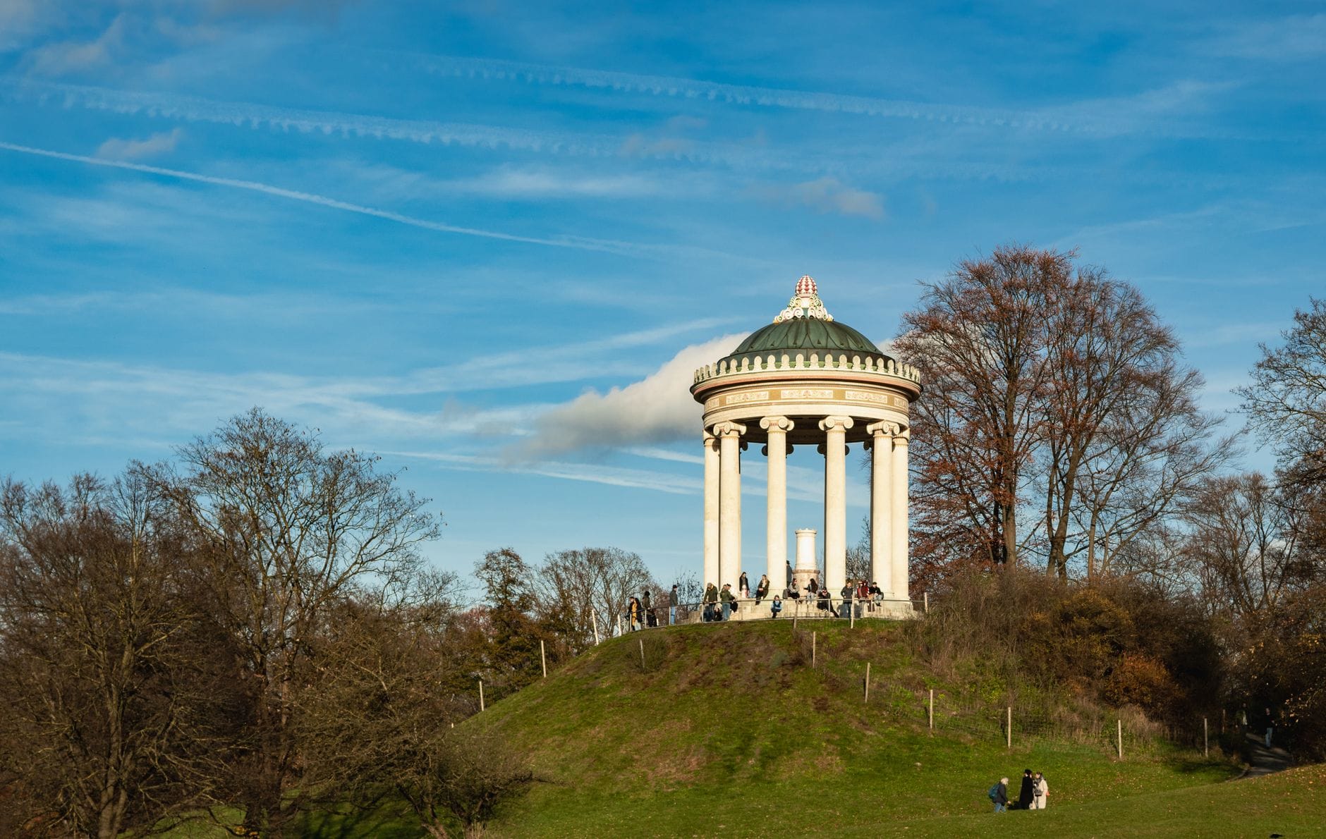 ผู้คนพักผ่อนบนเนินหญ้าบริเวณรอบวิหาร Monopteros ใน Englischer Garten เมืองมิวนิก โดยมีท้องฟ้าสีฟ้าและต้นไม้เป็นฉากหลัง
