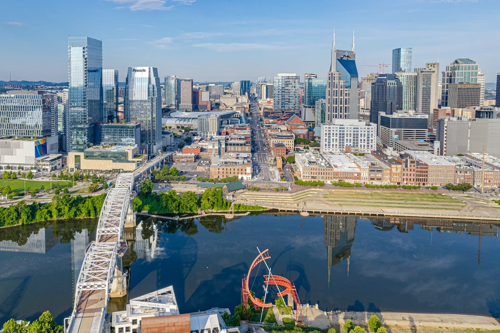 Vista aérea del horizonte del centro de Nashville con parques verdes, puente peatonal y río en primer plano en un día soleado.
