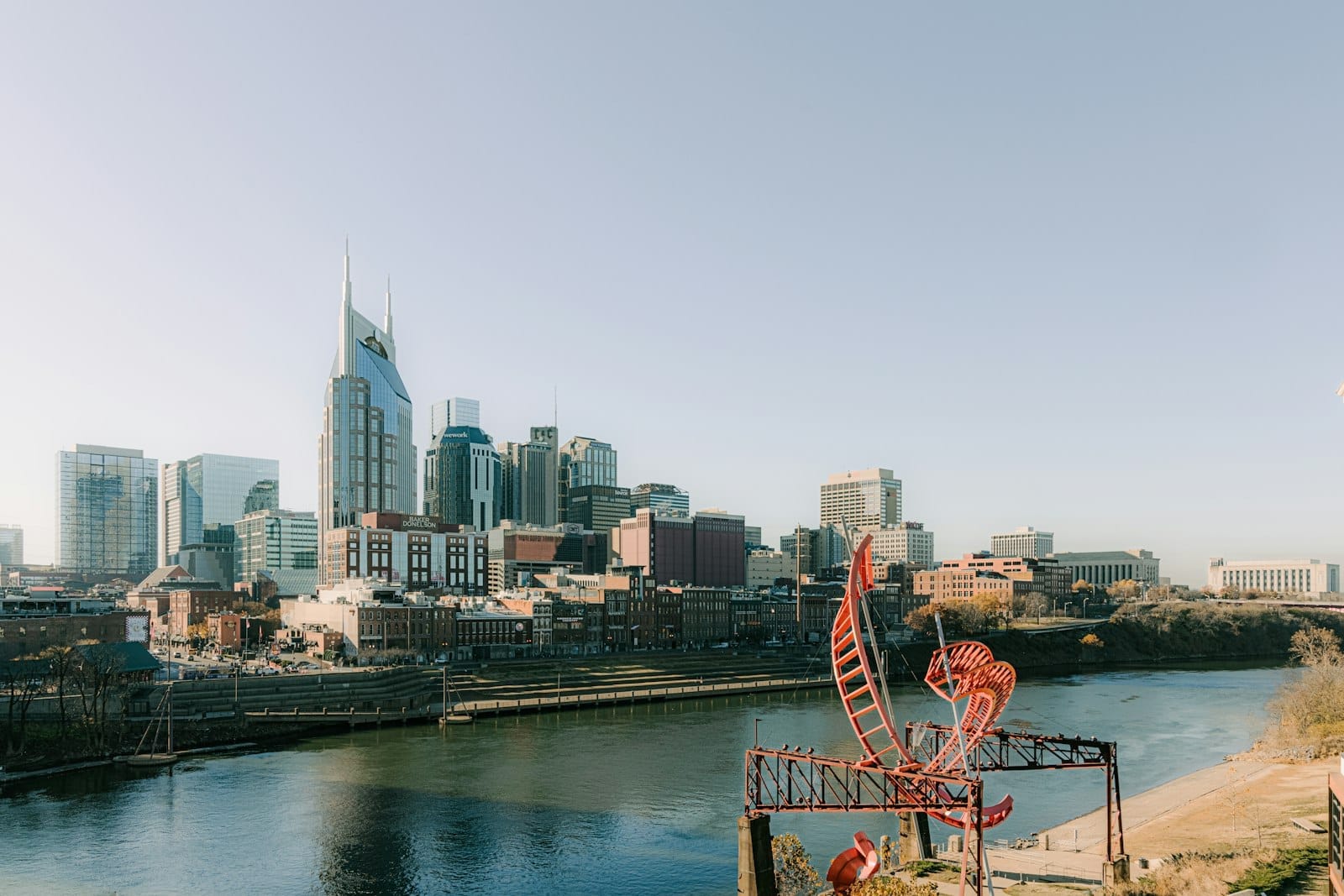 Vista panorámica del horizonte de Nashville desde el otro lado del río bajo un cielo azul despejado, con edificios modernos y una escultura roja artística en primer plano.