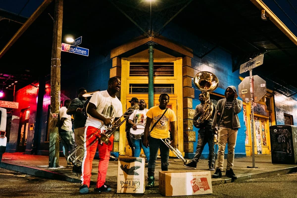 Eine Live-Brassband spielt nachts an einer berühmten Straßenecke in New Orleans unter Straßenschildern, mit bunten Gebäuden im Hintergrund.