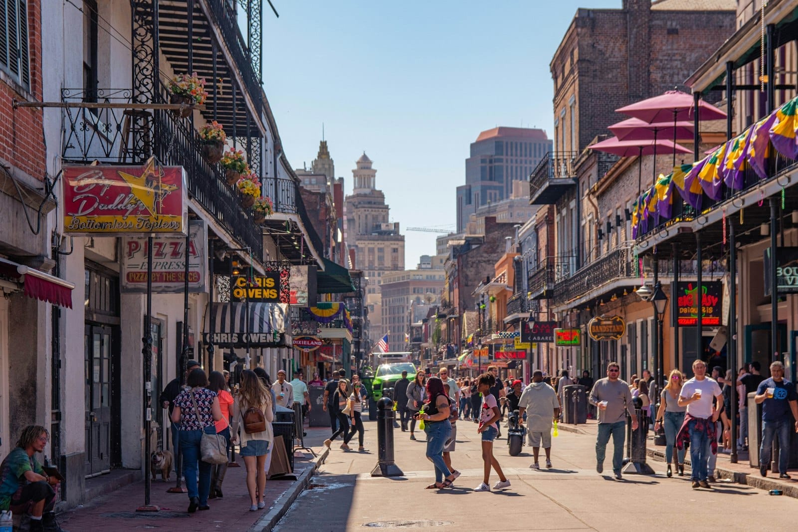 Pessoas caminhando por uma rua animada no Bairro Francês de Nova Orleans, com prédios históricos, sacadas e decorações coloridas do Mardi Gras.