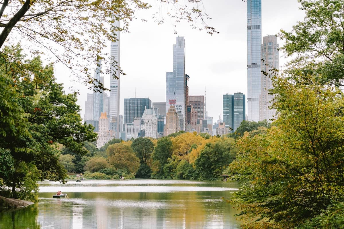 Blick auf den See des Central Park mit grünen Bäumen und Manhattan-Wolkenkratzern im Hintergrund, der die Außenbereiche von NYC an einem bewölkten Tag einfängt.
