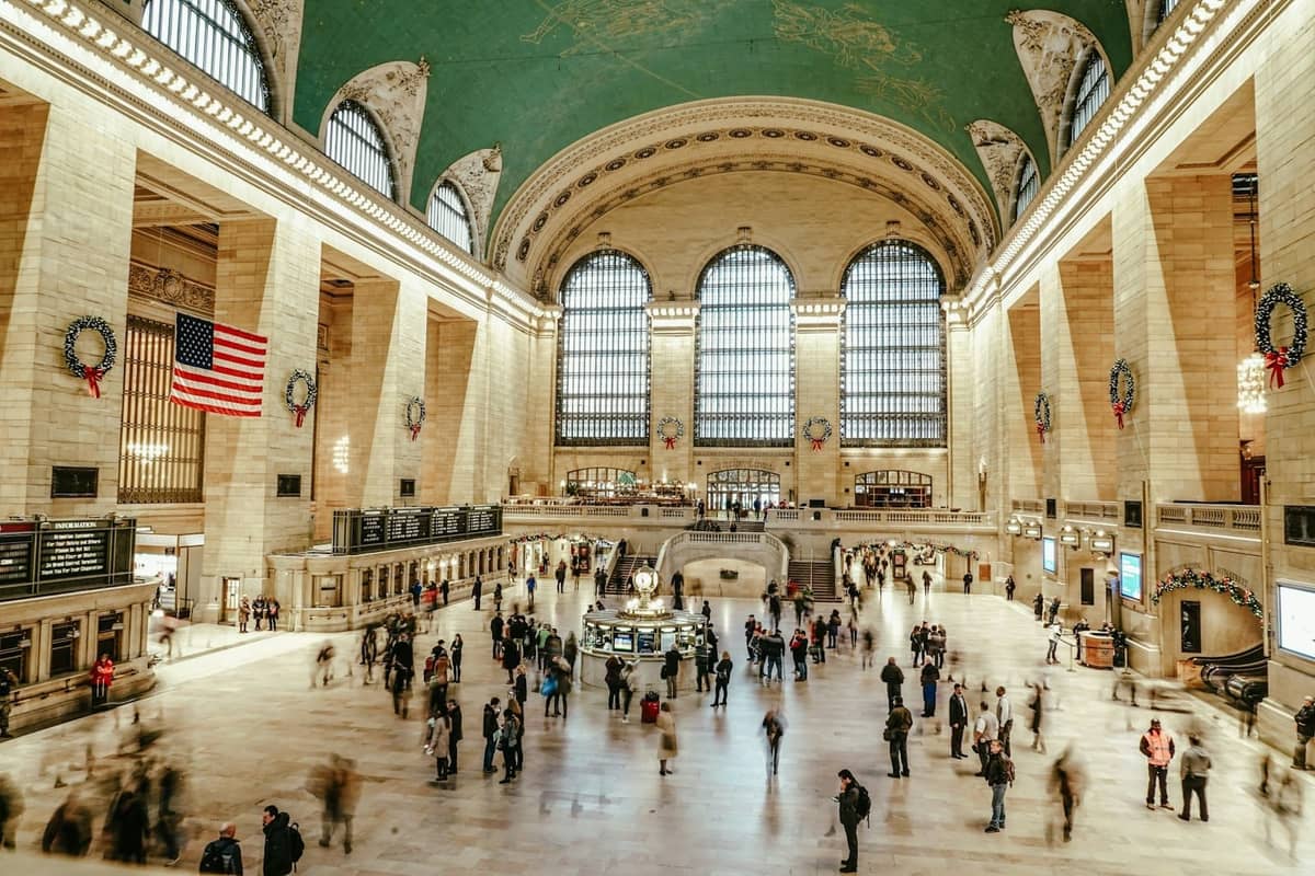 Die Haupthalle des Grand Central Terminal voller Menschen, mit ikonischen Bogenfenstern, der himmlischen Decke und der amerikanischen Flagge.