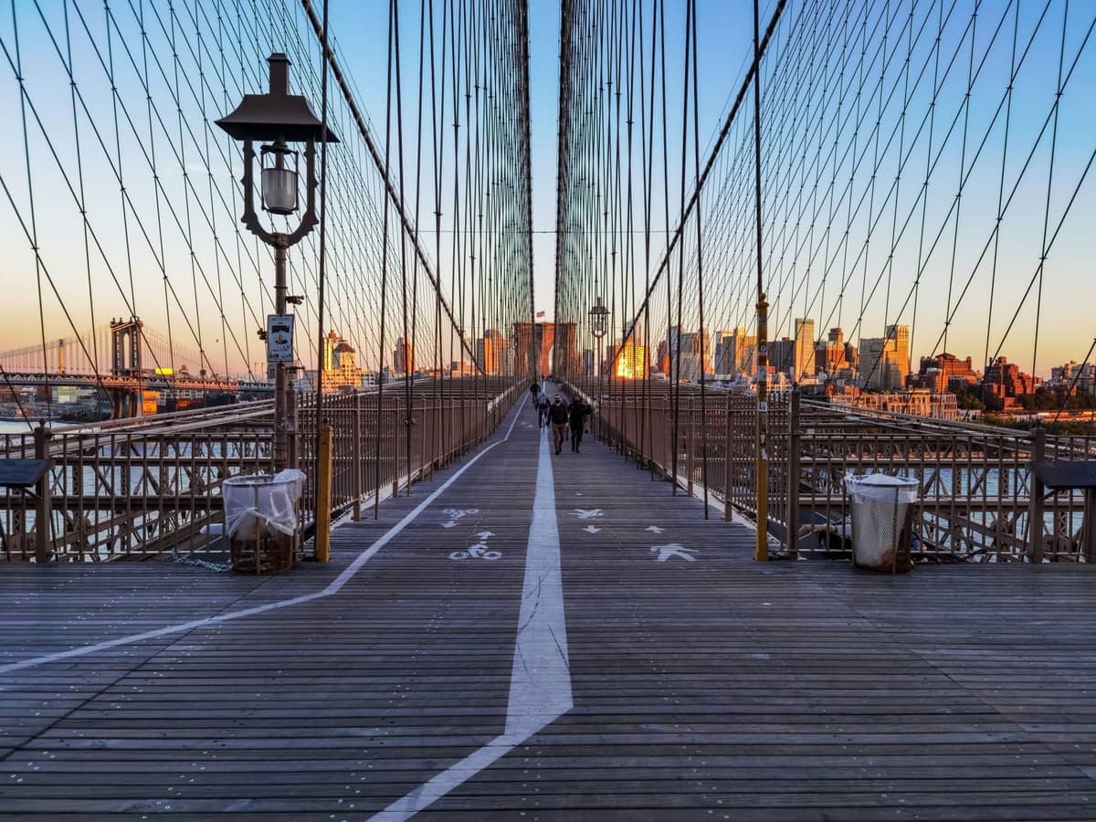 Fußgänger auf der Brooklyn Bridge mit Blick auf die Manhattan-Skyline bei Sonnenuntergang, ein klassisches kostenloses Erlebnis in New York City.