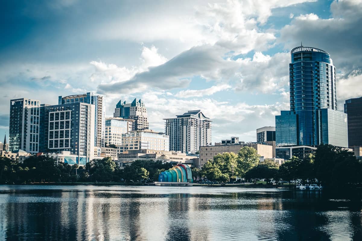 Vista dello skyline di Orlando con grattacieli accanto a un lago e le iconiche bande arcobaleno del Lake Eola visibili, sotto un cielo parzialmente nuvoloso.