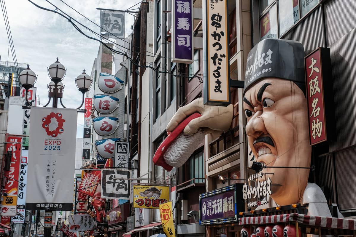 Bustling Dotonbori street in Osaka with colorful 3D restaurant signs, lanterns, banners, and crowds of people in daylight.