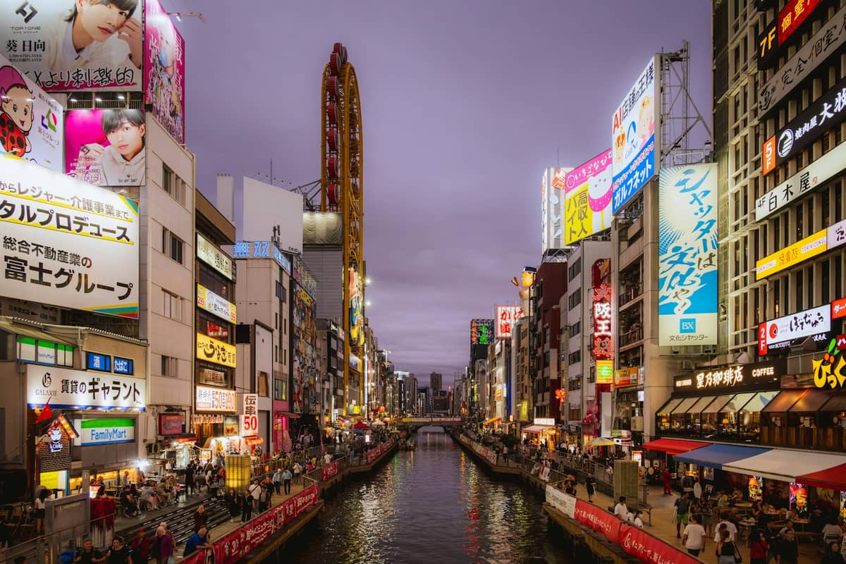 Wide view of Osaka's iconic Dotonbori canal with crowds, neon signs, bright billboards, and busy walkways lining both sides at dusk.