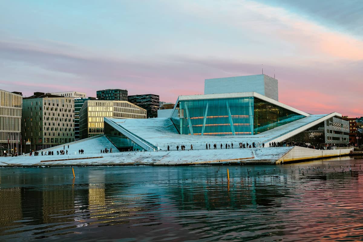 Oslo Opera House พร้อมผู้คนเดินบนหลังคาลาดเอียงยามพระอาทิตย์ตก มีอาคารสมัยใหม่และน้ำอยู่เบื้องหน้า