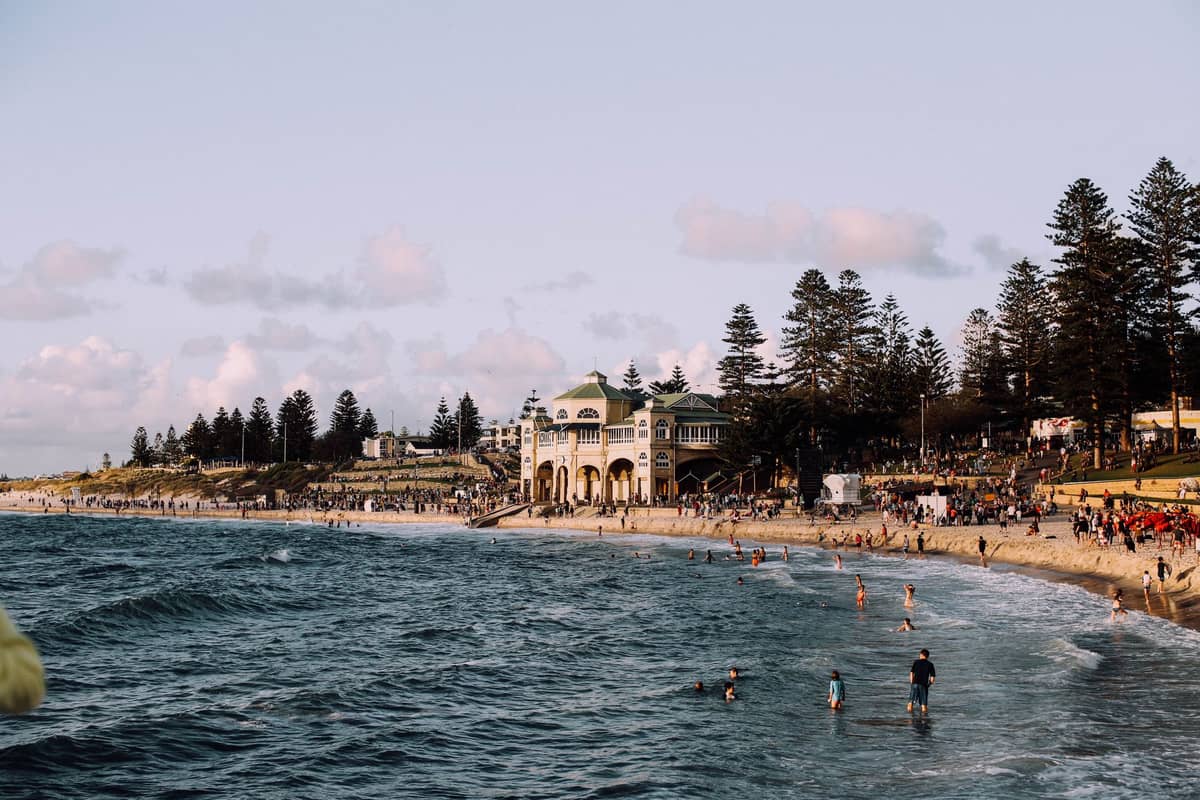 Orilla concurrida en Cottesloe Beach en Perth con bañistas, olas, pinos y el distintivo pabellón histórico.