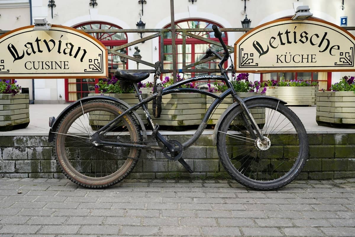 Altes schwarzes Fahrrad vor dem Schild eines lettischen Küche-Restaurants geparkt, mit Blumenkübeln und roten Bogenfenstern im Hintergrund.