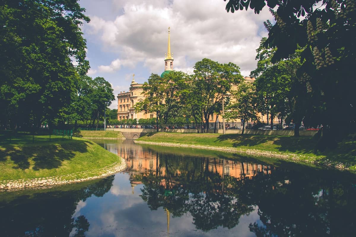 Ein malerischer Blick auf einen von Bäumen gesäumten Kanal in Riga mit grünem Gras, spiegelndem Wasser und einem historischen gelben Gebäude im Hintergrund.
