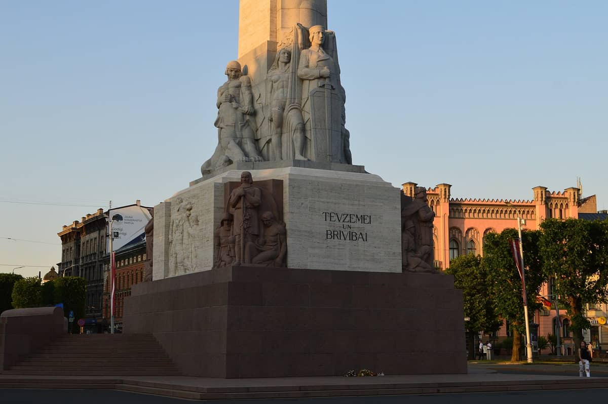Weitwinkelansicht der Basis des Freiheitsdenkmals in Riga mit detaillierten Statuen, eingraviertem Text und der städtischen Umgebung bei Sonnenuntergang.