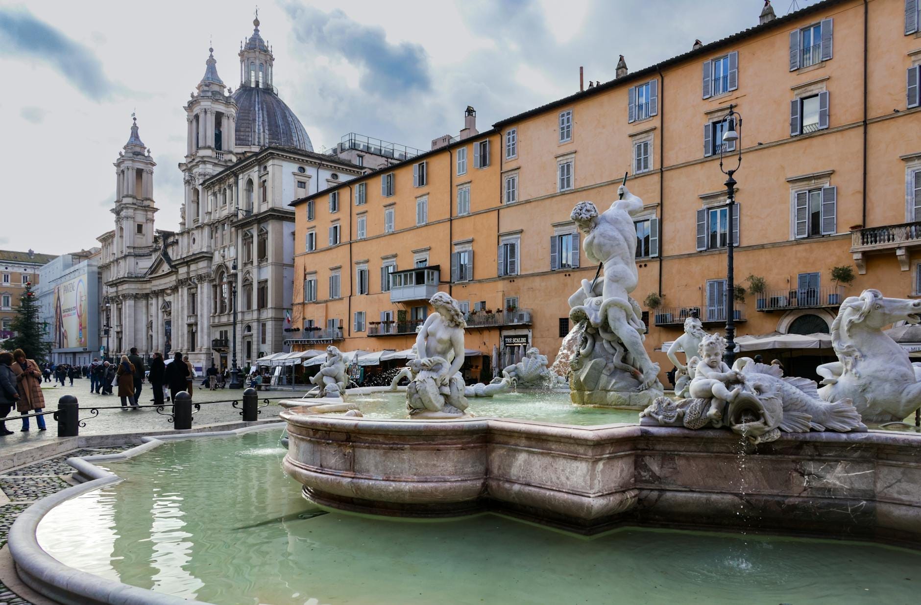 Baroque fountain with elaborate statues in Piazza Navona, Rome surrounded by historic buildings and Sant'Agnese in Agone church in the background.