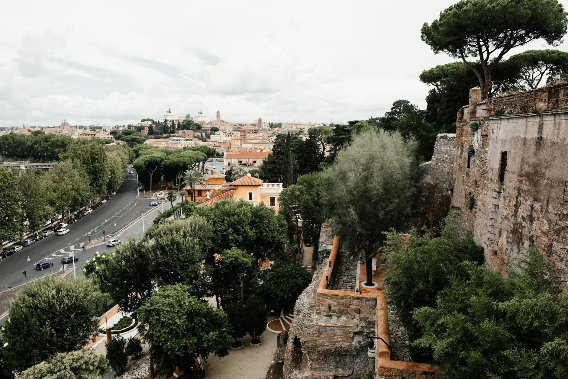 View from a hilltop in Rome with cityscape, trees, ancient stone walls, and a road winding through greenery.