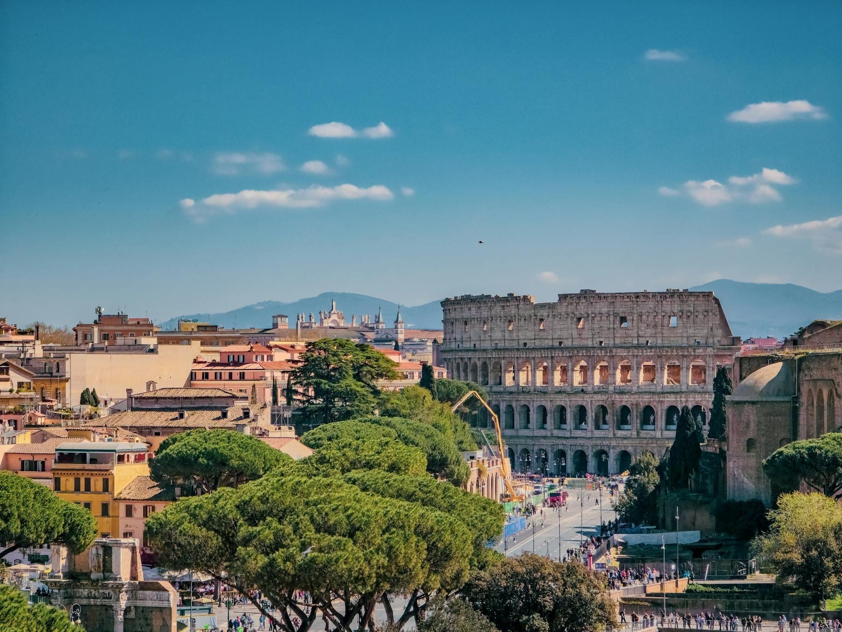 Panoramic view of Rome featuring the Colosseum, city rooftops, trees, and distant mountains under a blue sky with scattered clouds.