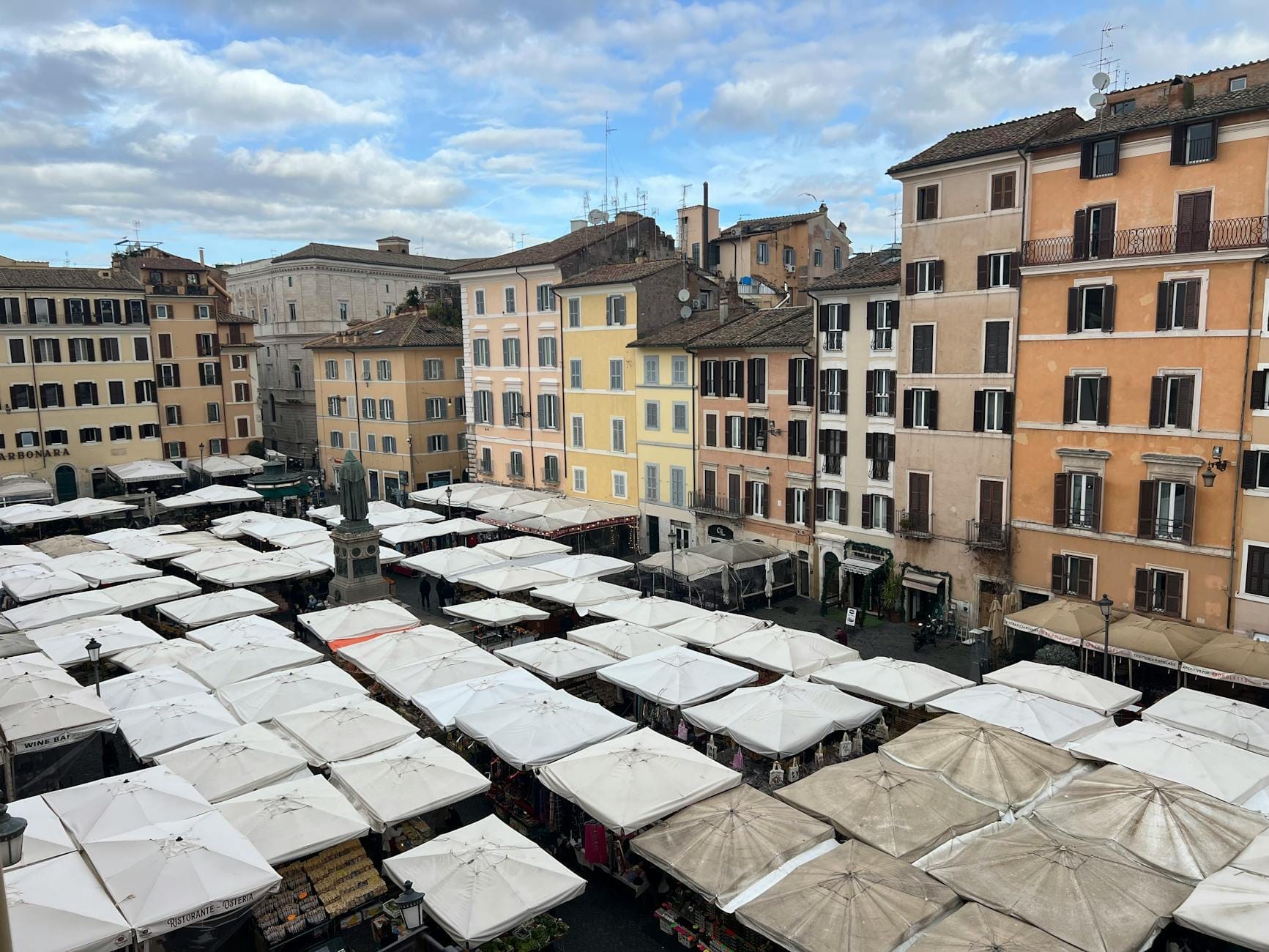 A vibrant Roman square with dozens of market stalls under white canopies, surrounded by colorful old apartment buildings and open sky.