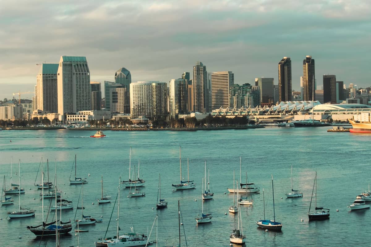 Die Skyline von San Diego bei Sonnenuntergang, vom anderen Ufer der Bucht aus gesehen, mit ruhigem Wasser und Segelbooten im Vordergrund.