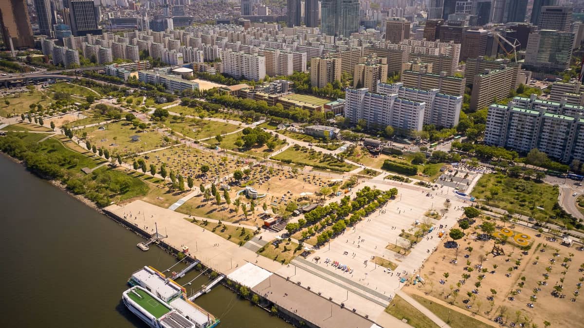 Vista aérea de um grande parque à beira do rio em Seul com trilhas, áreas verdes e apartamentos de alto padrão nas proximidades.