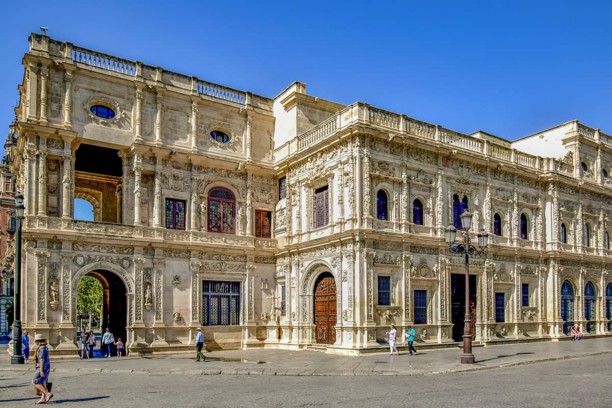 อาคารประวัติศาสตร์อันยิ่งใหญ่และประดับประดาอย่างวิจิตรในเมือง Seville พร้อมงานหินแกะสลักละเอียด ประตูโค้งขนาดใหญ่ และผู้คนเดินอยู่ด้านนอกในวันที่แดดจ้า