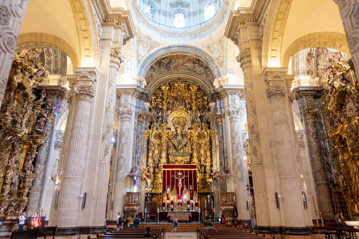 ภาพกว้างของภายในโบสถ์บาโรกที่ประดับประดาอย่างวิจิตรในเมือง Seville พร้อมเสาสูง แท่นบูชาสีทอง การตกแต่งที่ซับซ้อน และนักท่องเที่ยวในที่นั่ง