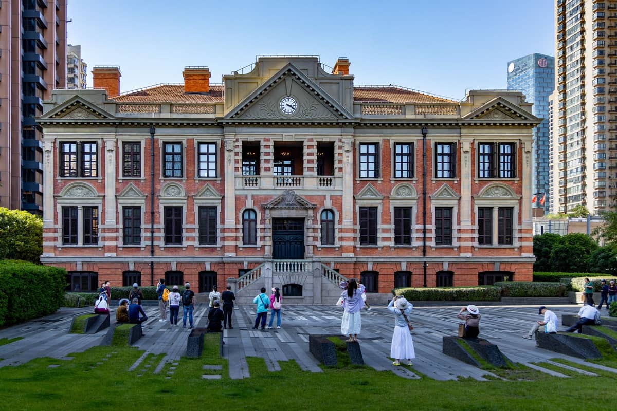 Bâtiment historique en brique avec une horloge au-dessus de l'entrée, des personnes rassemblées devant, entouré de gratte-ciel modernes à Shanghai.