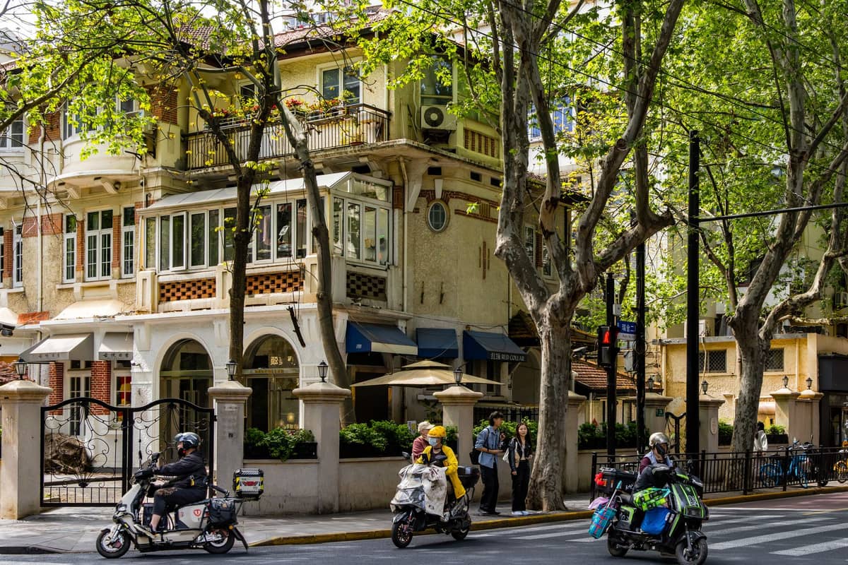 Coin de rue historique de la Concession Française à Shanghai avec de grands platanes, un bâtiment Art Déco et des personnes à scooter et à vélo.