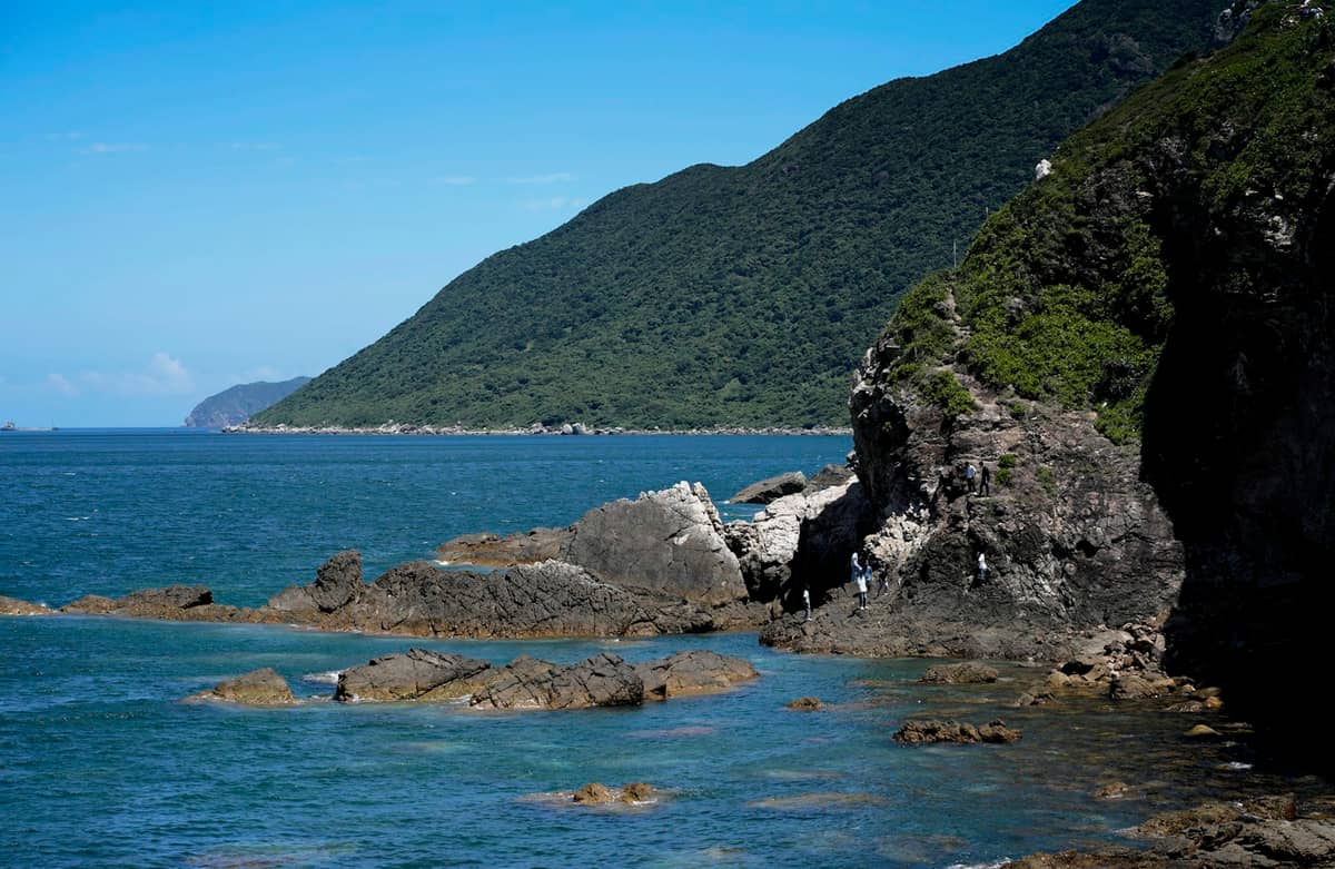 Costa rochosa com colinas verdes exuberantes encontrando o oceano azul sob um céu limpo, evocando as belas áreas de caminhada costeira de Shenzhen.