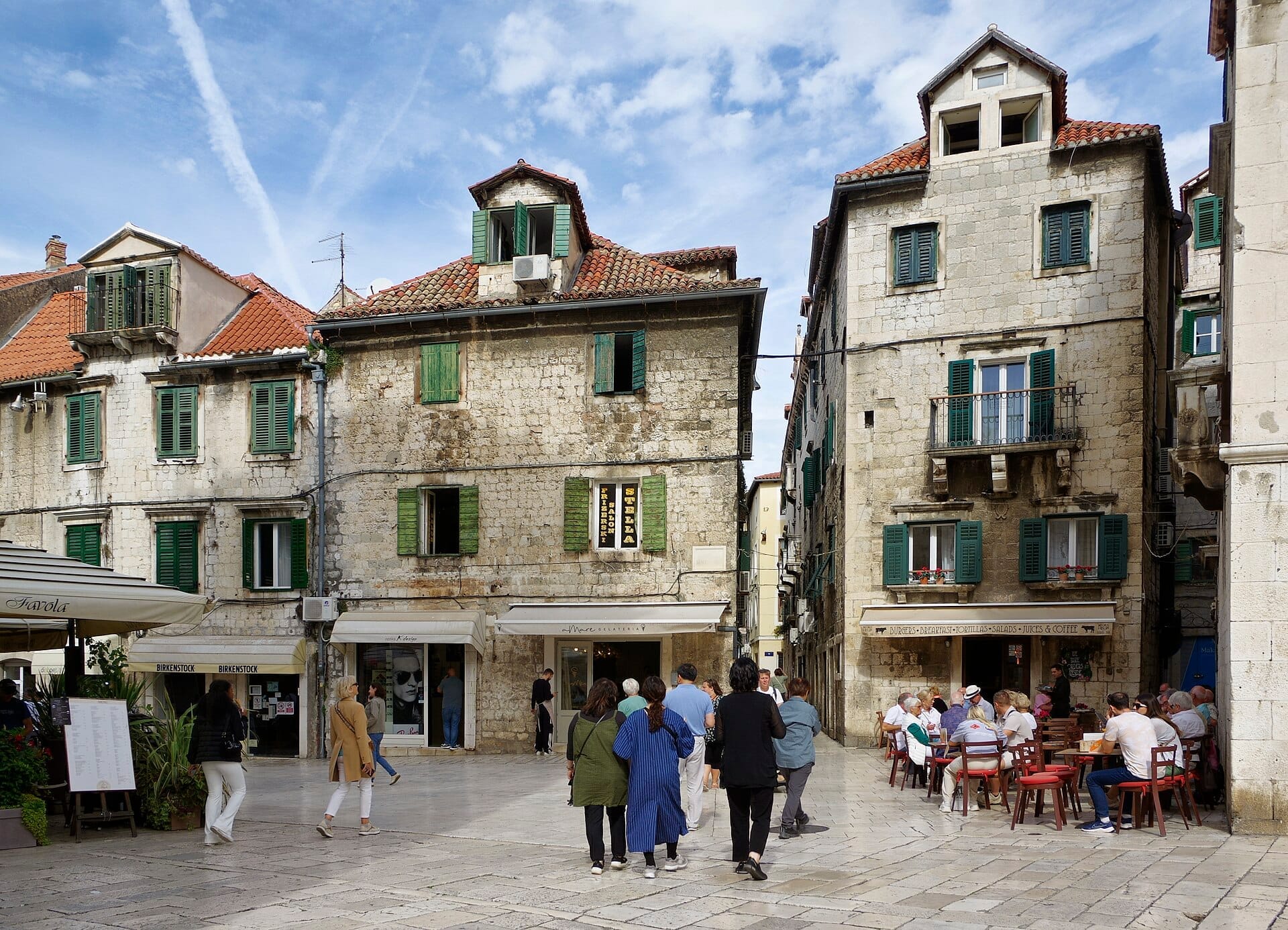 Pessoas passeiam por uma praça ensolarada na cidade antiga de Split, rodeada de edifícios de pedra com venezianas verdes e telhados de terracota.