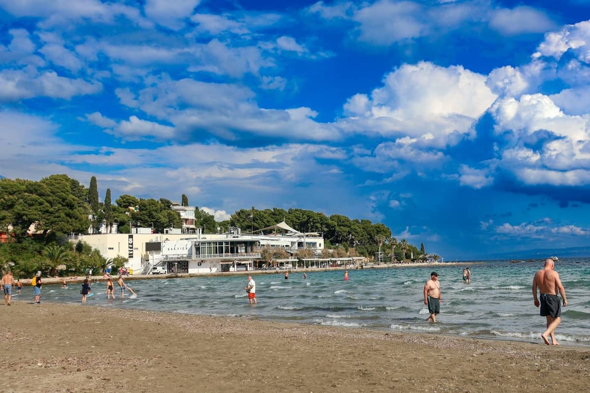 Persone che guadano e si rilassano su una spiaggia sabbiosa a Spalato, in Croazia, con edifici, alberi e un vivace cielo azzurro sullo sfondo.