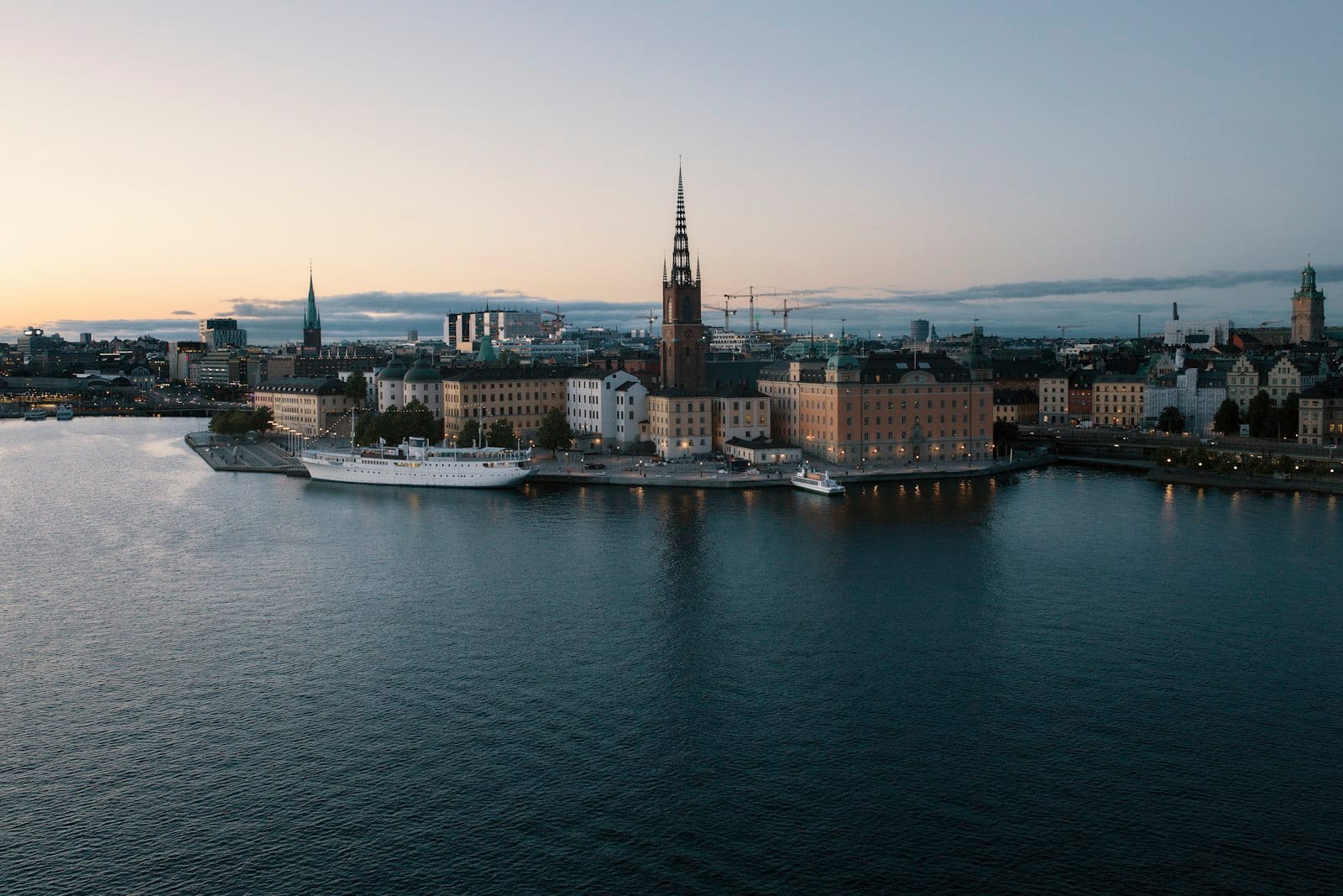 ภาพมุมสูงของเกาะ Riddarholmen ในสตอกโฮล์ม เห็นยอดโบสถ์ Riddarholmskyrkan ตึกโบราณริมน้ำ เรือ และทิวทัศน์เมืองยามเย็น น้ำสงบล้อมรอบ