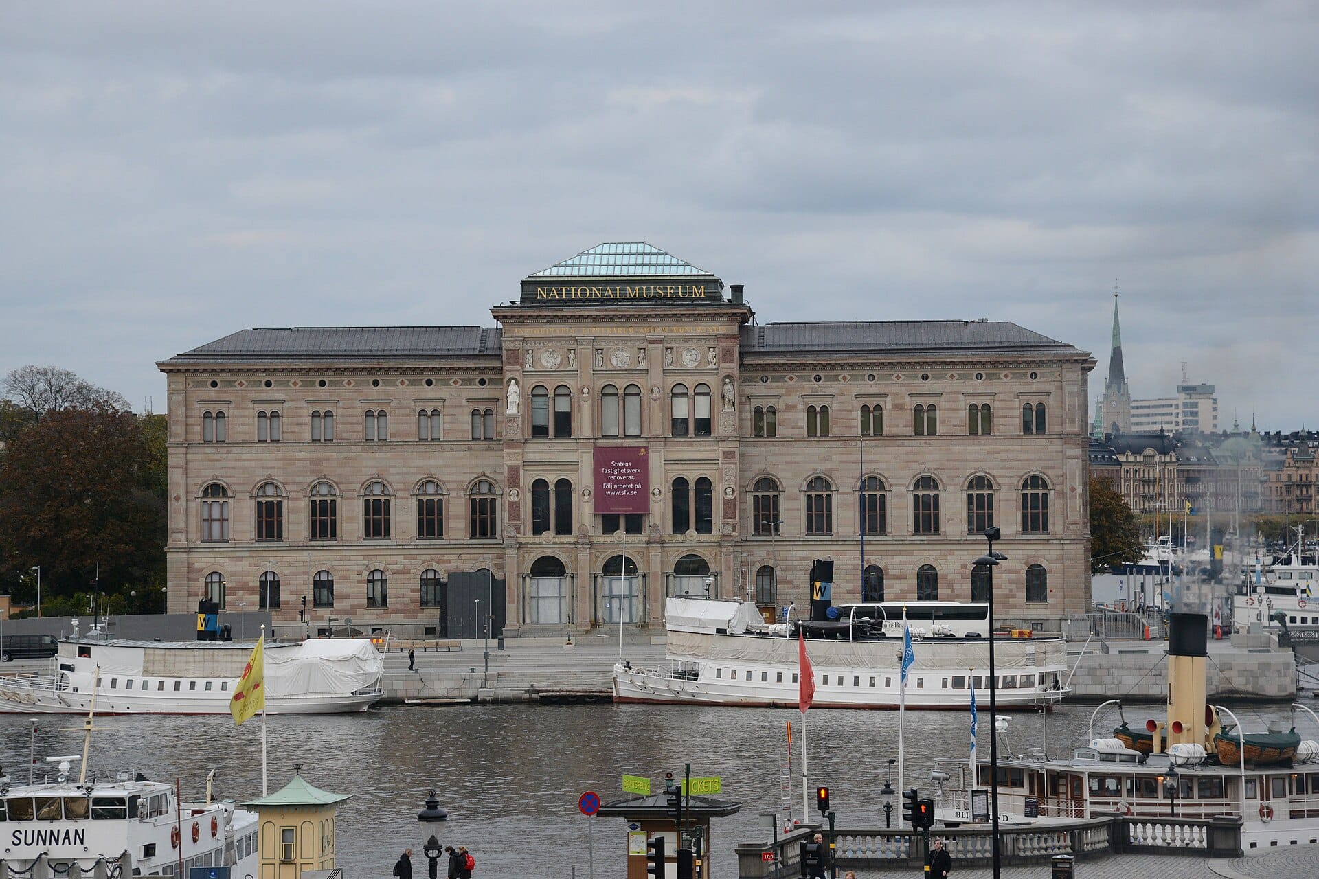 ด้านหน้าของ Nationalmuseum ในสตอกโฮล์มที่มองเห็นข้ามน้ำ โดยมีเรือและอาคารในเมืองอยู่เบื้องหน้าในวันที่มีเมฆมาก
