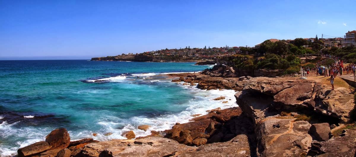 Scenic Sydney coastal walk along cliffs with turquoise sea, swimmers, and crowds walking on a path, backed by urban eastern suburbs.